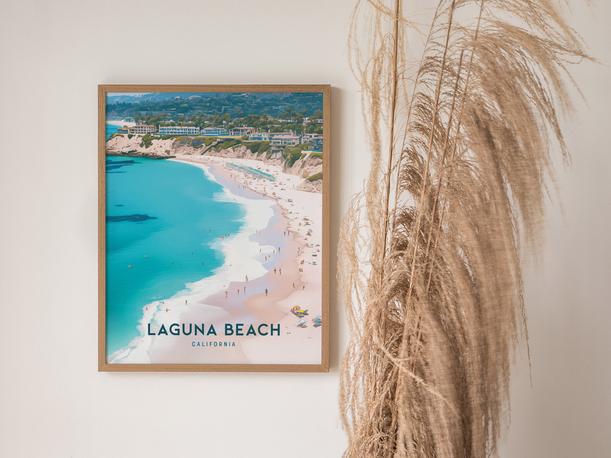 A framed poster of Laguna Beach, California, featuring a coastline with turquoise water and sandy shores, is displayed on a wall beside tall, dried pampas grass.