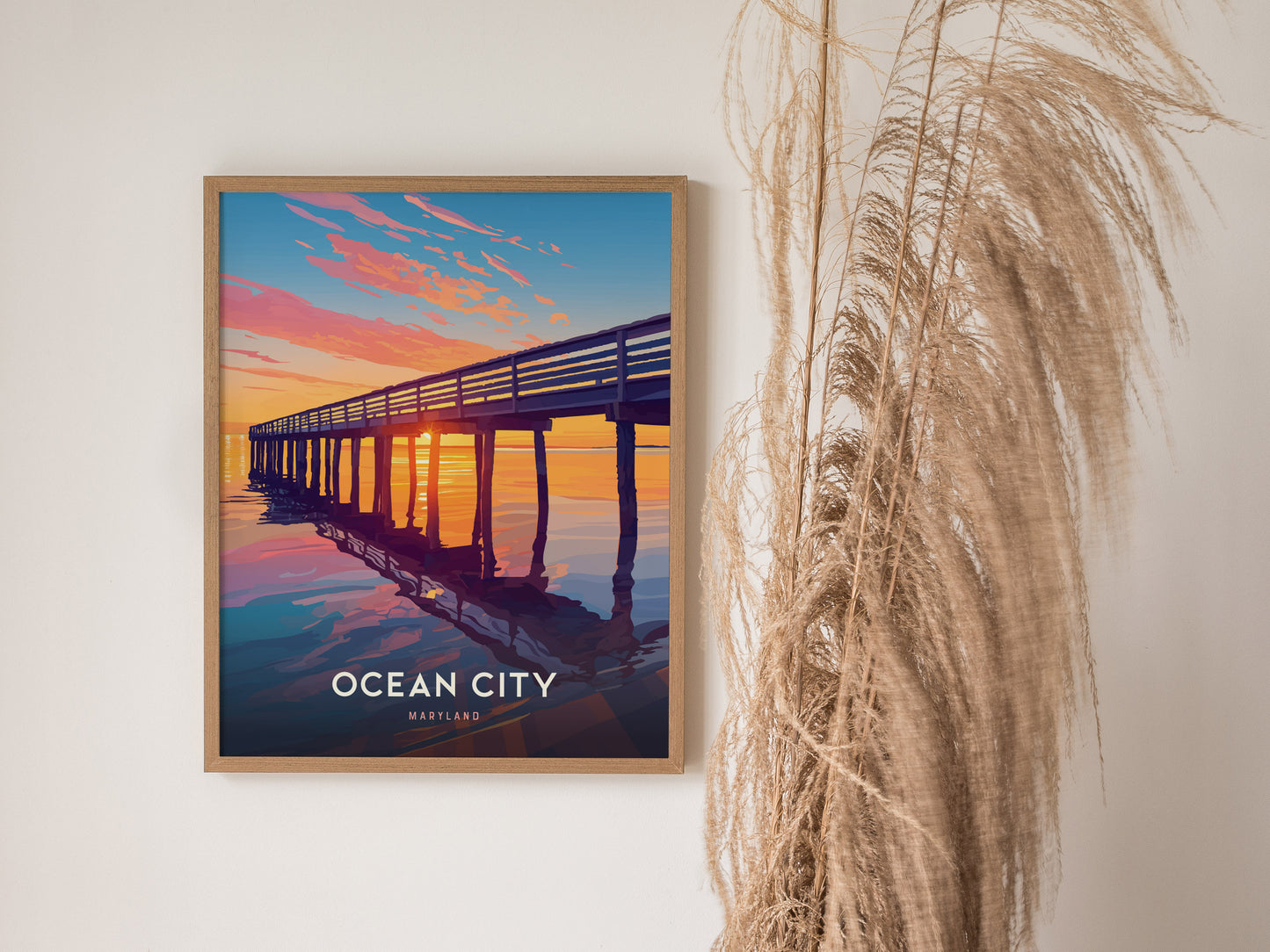 A framed poster of Ocean City, Maryland, featuring a vibrant sunset over a pier, hangs on a white wall beside decorative dried grasses.