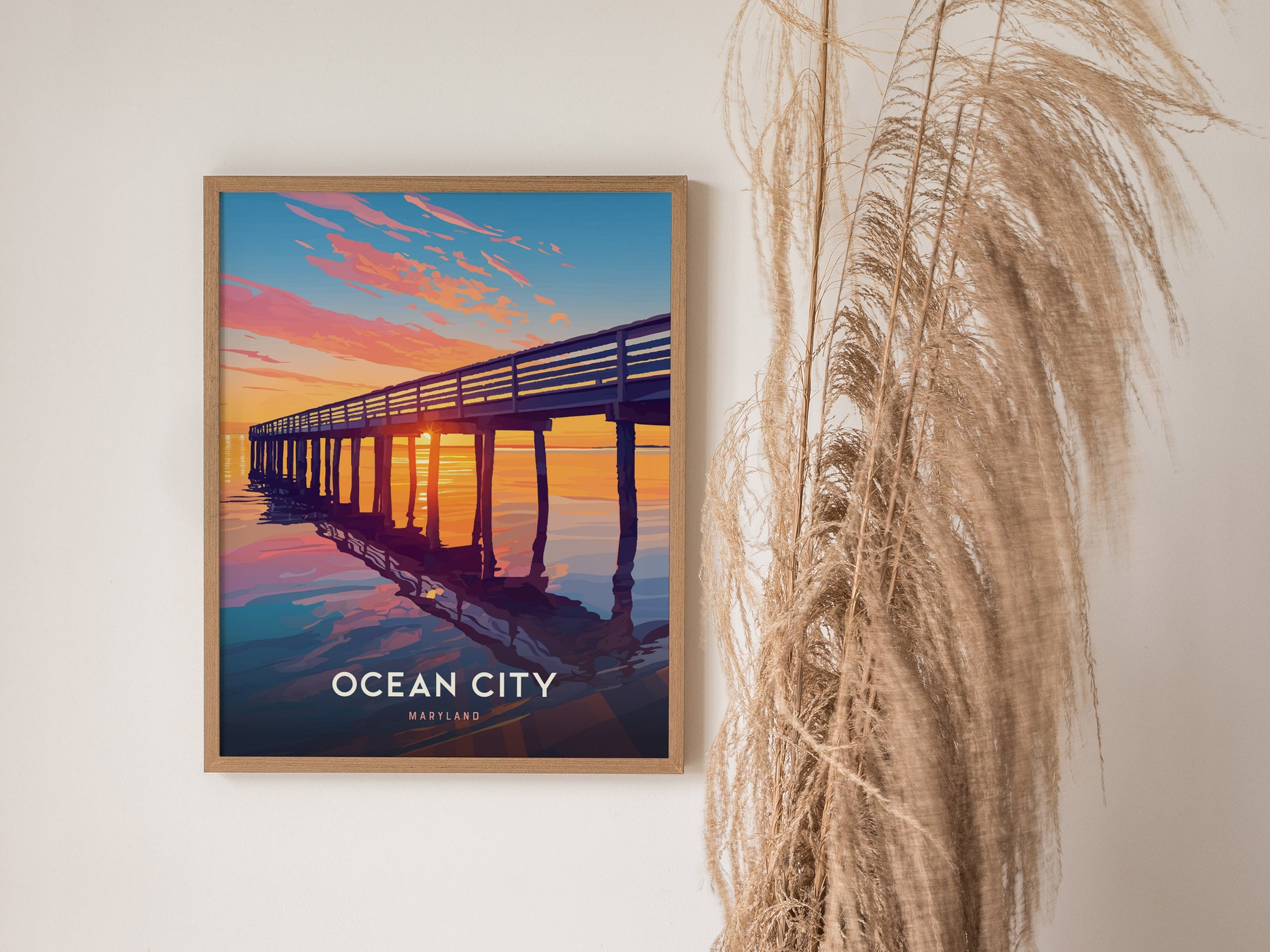 A framed poster of Ocean City, Maryland, featuring a vibrant sunset over a pier, hangs on a white wall beside decorative dried grasses.
