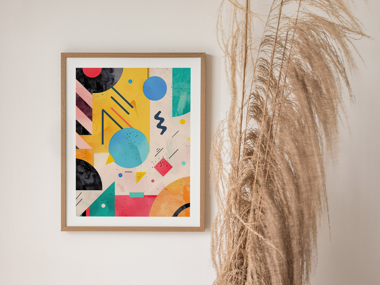 A framed abstract poster featuring vibrant geometric shapes and bold colors hangs on a white wall next to tall, dried pampas grass.