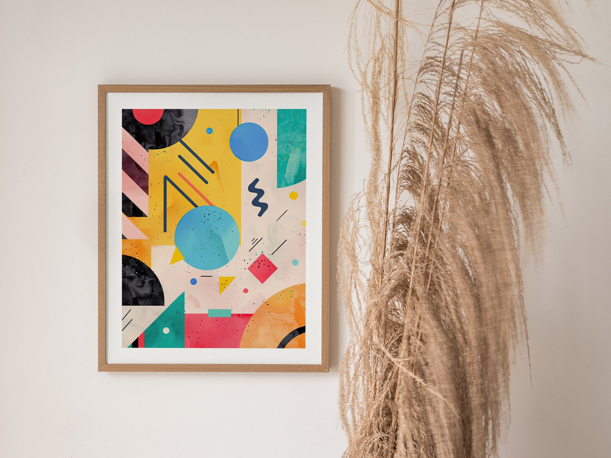 A framed abstract poster featuring vibrant geometric shapes and bold colors hangs on a white wall next to tall, dried pampas grass.