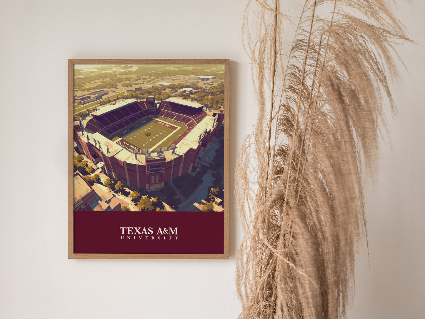 A framed poster of Kyle Field at Texas A&M University hangs on a white wall beside tall, dried grass. The artwork features the stadiums aerial view with bold colors.