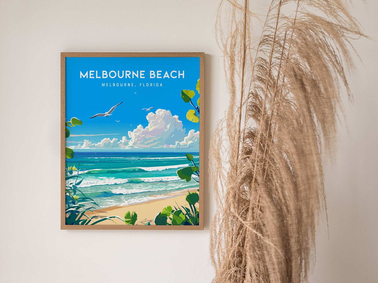 A framed poster of Melbourne Beach, Florida, features a vibrant seascape with blue skies, ocean waves, and seagulls. Its displayed next to tall, dried grasses for a coastal decor vibe.