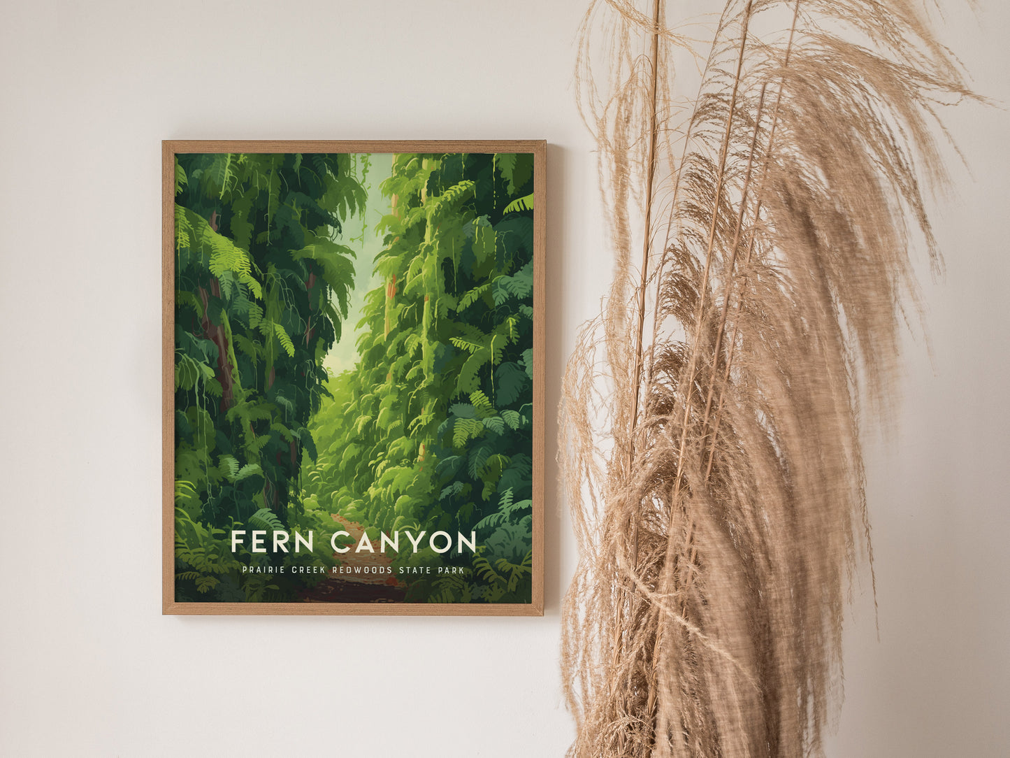 A framed poster of Fern Canyon hangs on a white wall, featuring lush green foliage. Tall, dried grass decorates the space beside the artwork.