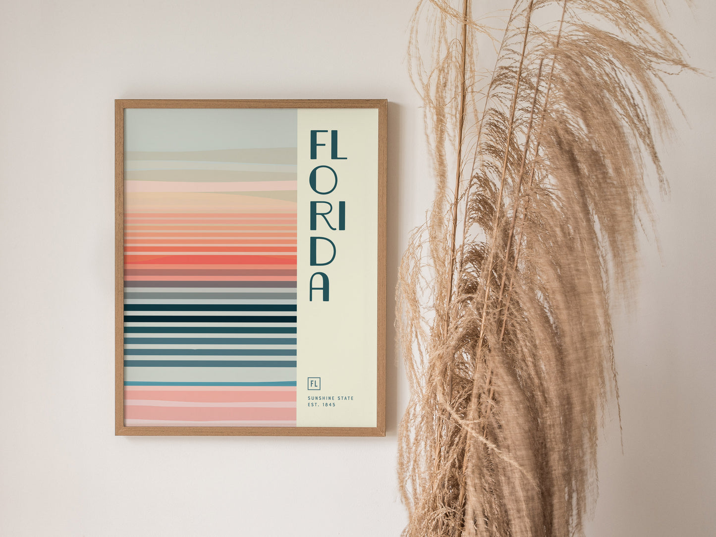 A framed abstract poster featuring horizontal pastel stripes and the word FLORIDA hangs on a white wall beside dried pampas grass. The design is minimalistic and serene.