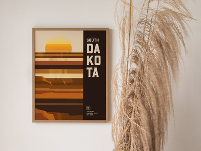 Framed South Dakota poster features a minimalist geometric design with earthy tones and a stylized sunset. It hangs beside tall, dried pampas grass against a white wall.
