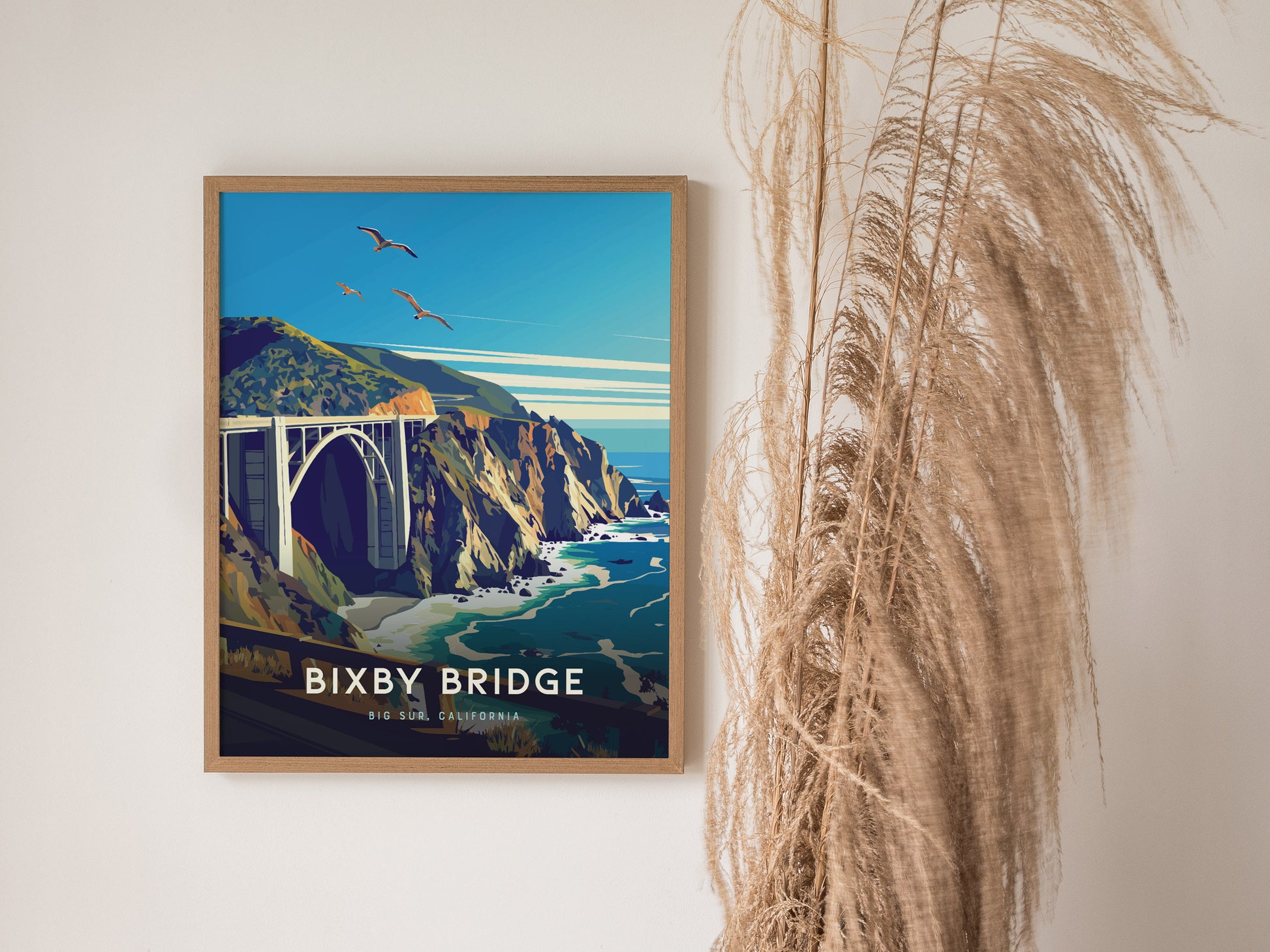 A framed poster of Bixby Bridge in Big Sur, California, depicts the structure against rugged cliffs and turquoise waters, with birds flying above, placed beside decorative dried grasses.
