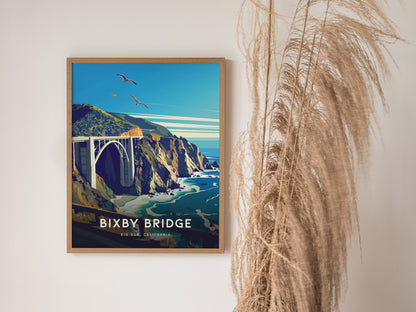 A framed poster of Bixby Bridge in Big Sur, California, depicts the structure against rugged cliffs and turquoise waters, with birds flying above, placed beside decorative dried grasses.