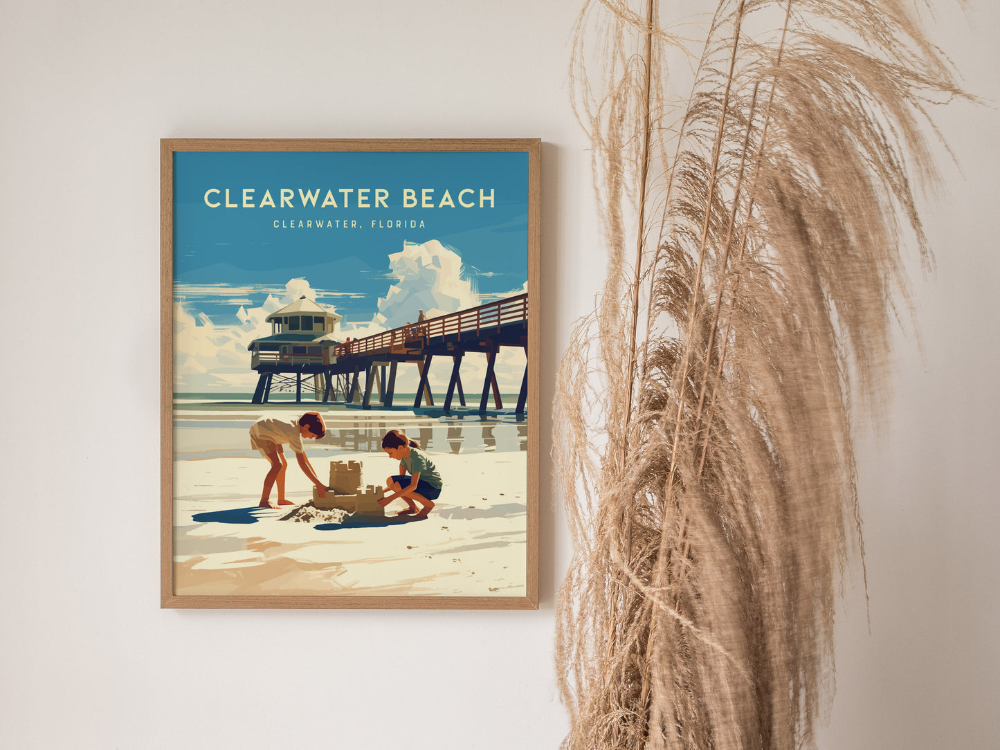 Framed poster of Clearwater Beach showing two children building a sandcastle near a pier under a blue sky. Tall, dried grasses are visible beside the artwork.