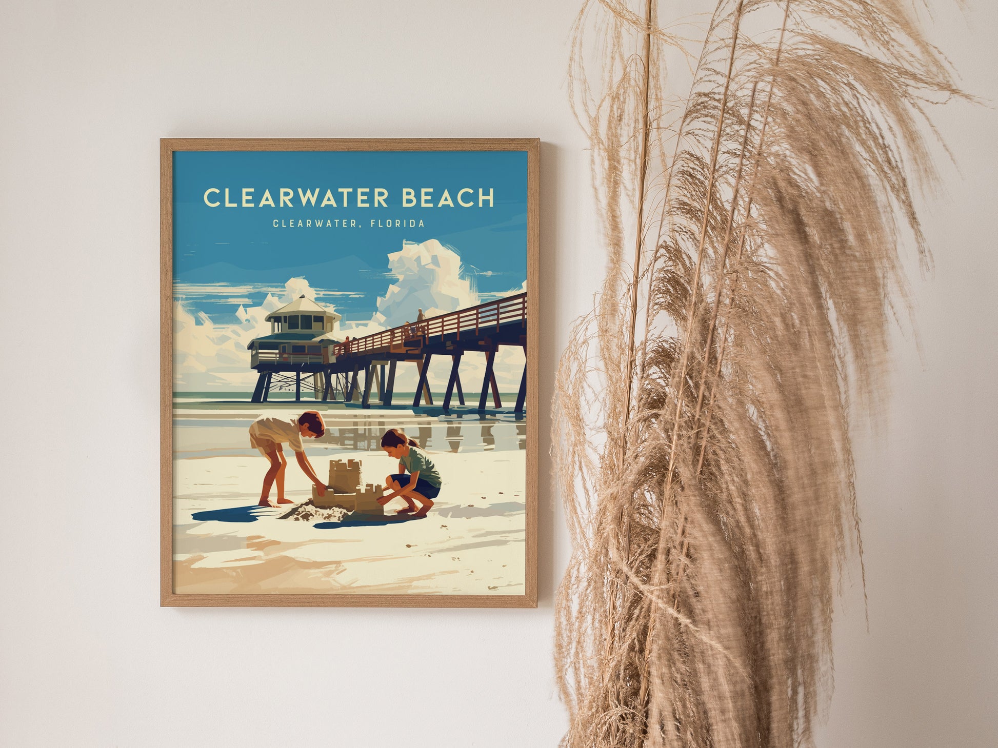 Framed poster of Clearwater Beach showing two children building a sandcastle near a pier under a blue sky. Tall, dried grasses are visible beside the artwork.