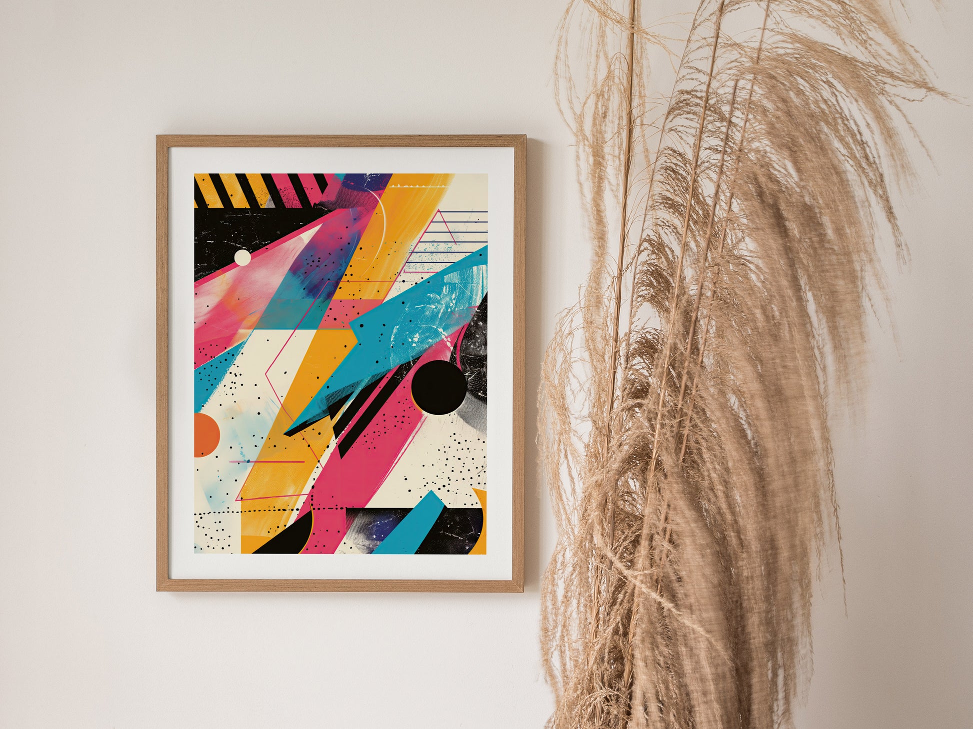 A framed poster featuring vibrant 1980s-style geometric shapes and colors is displayed on a white wall next to tall, dried pampas grass.