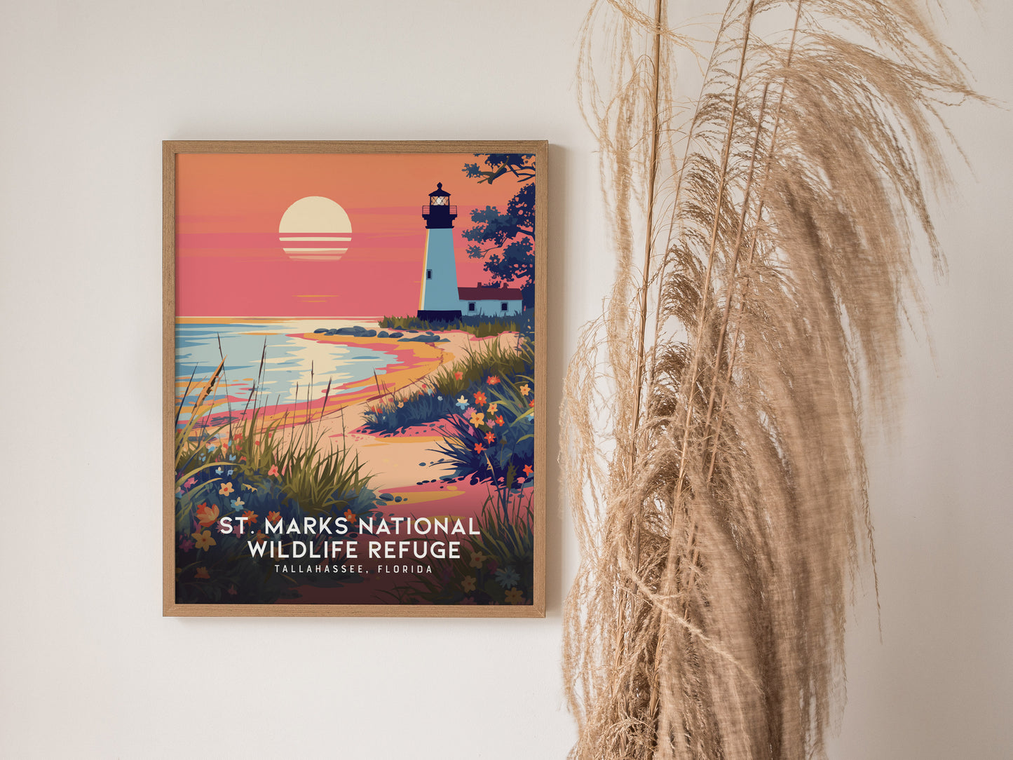 A framed poster of St. Marks National Wildlife Refuge featuring a lighthouse at sunset with vibrant coastal scenery is displayed on a wall beside tall dried grasses.