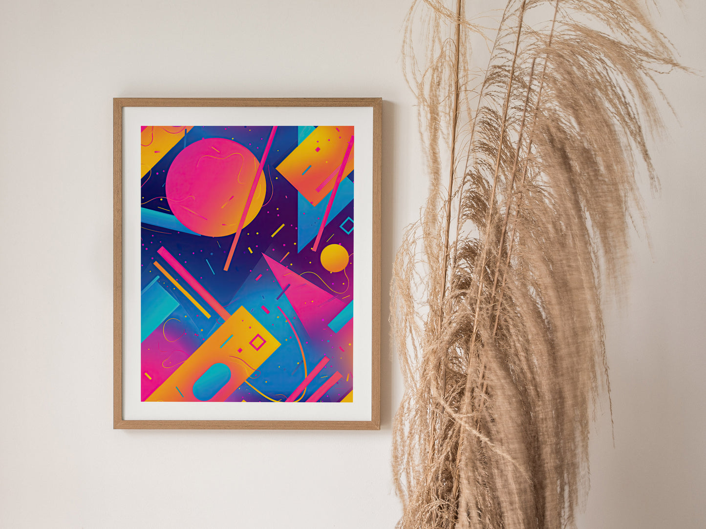 A framed poster featuring vibrant neon geometric shapes in pink, purple, yellow, and blue hangs on a wall next to tall, dried pampas grass.