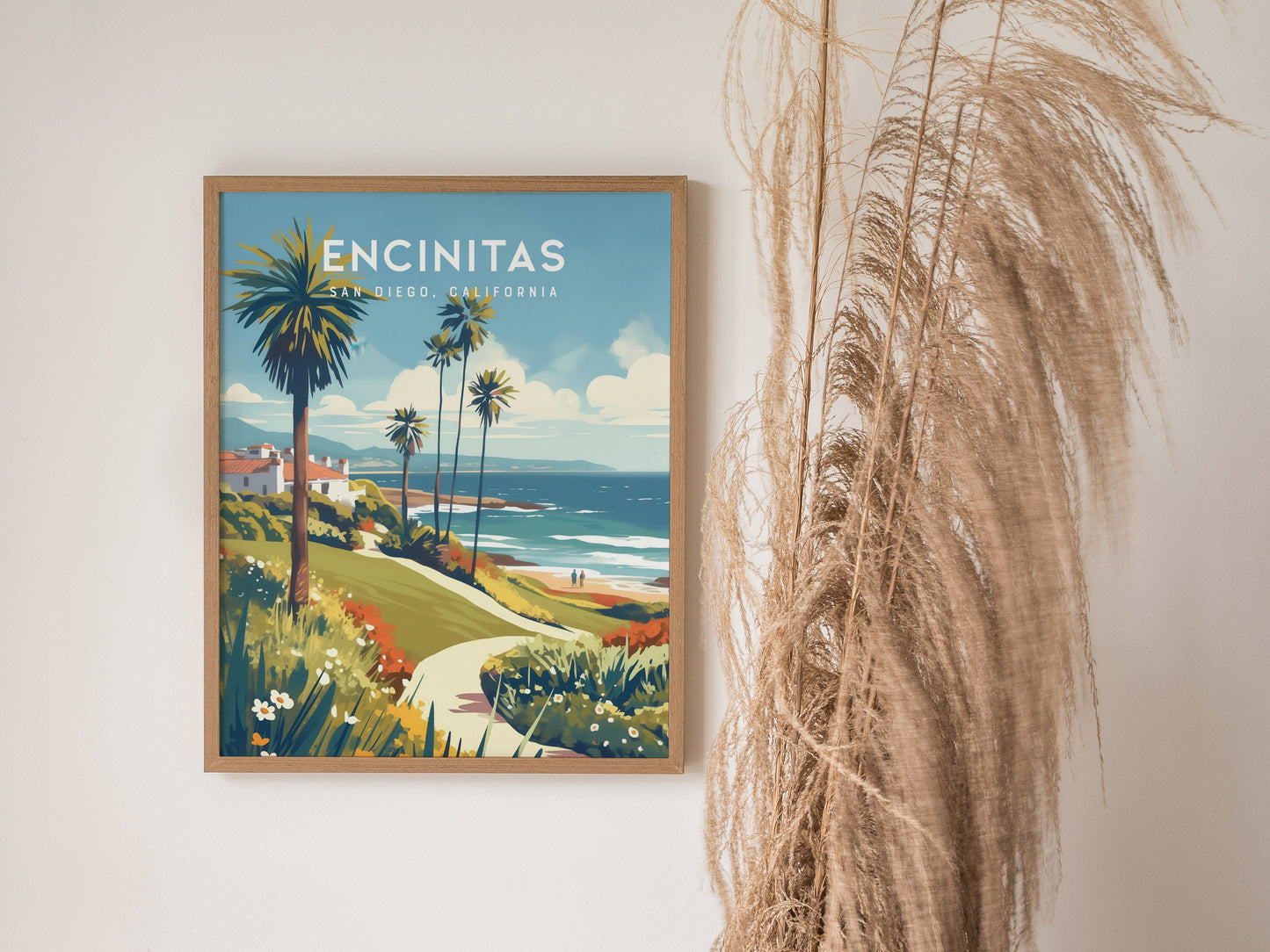 A framed poster of Encinitas, California, featuring a vibrant coastal scene with palm trees and ocean views, is displayed on a wall next to tall dried pampas grass.