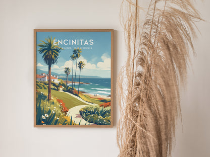 A framed poster of Encinitas, California, featuring a vibrant coastal scene with palm trees and ocean views, is displayed on a wall next to tall dried pampas grass.