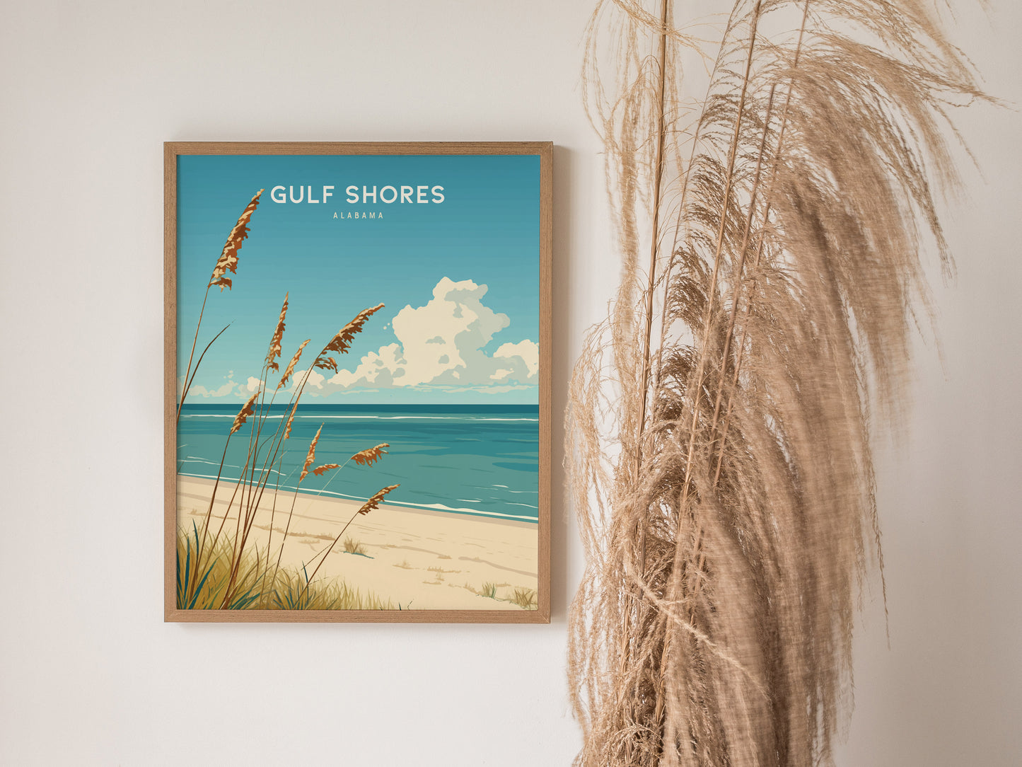 A framed Gulf Shores, Alabama poster depicts a serene beach scene with golden dunes and blue ocean, displayed next to dried pampas grass against a white wall.
