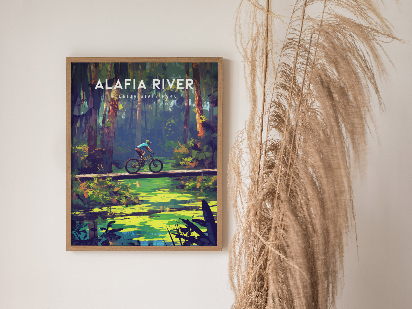 Framed poster of Alafia River State Park featuring a cyclist on a wooden bridge over a green creek, surrounded by lush forest. Dried grass decorates the right side.