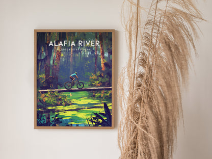 Framed poster of Alafia River State Park featuring a cyclist on a wooden bridge over a green creek, surrounded by lush forest. Dried grass decorates the right side.
