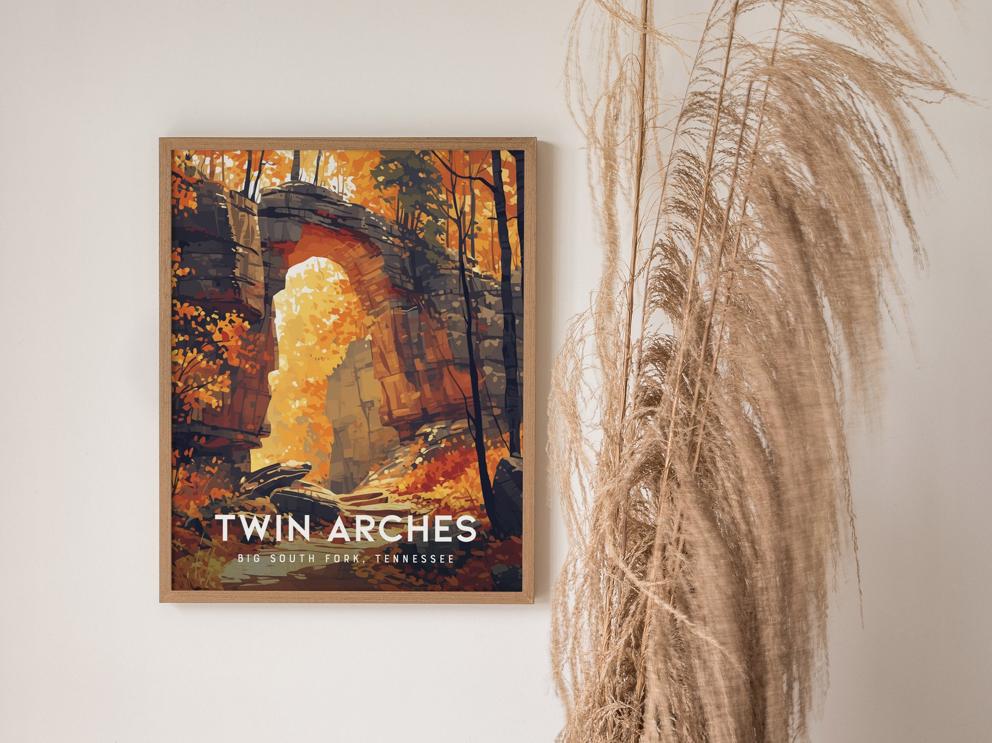 A framed poster of the Twin Arches in Big South Fork, Tennessee, features vibrant autumn foliage and sandstone formations. It is displayed next to decorative dried plants.