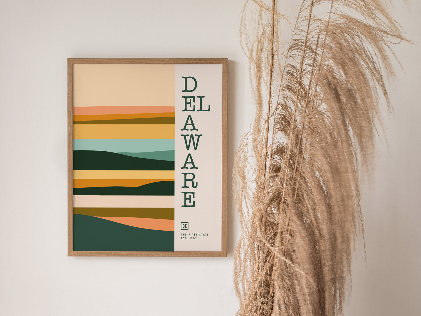A framed Delaware minimalist poster features abstract, layered shapes in earthy tones with vertical text. It hangs beside tall, dried pampas grass against a neutral wall.