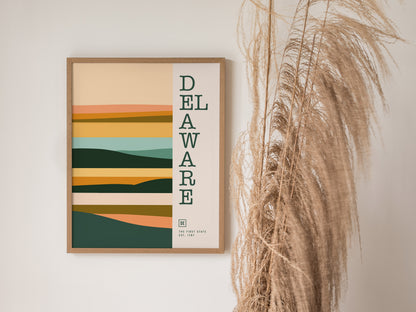 A framed Delaware minimalist poster features abstract, layered shapes in earthy tones with vertical text. It hangs beside tall, dried pampas grass against a neutral wall.
