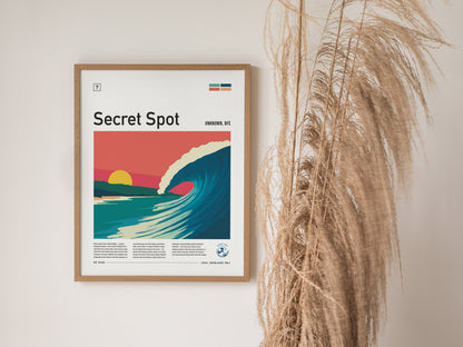 A framed poster titled Secret Spot features a vibrant illustration of a curling wave under a sunset with pastel colors. The artwork is accompanied by descriptive text and set against a neutral wall, complemented by decorative dried grass.