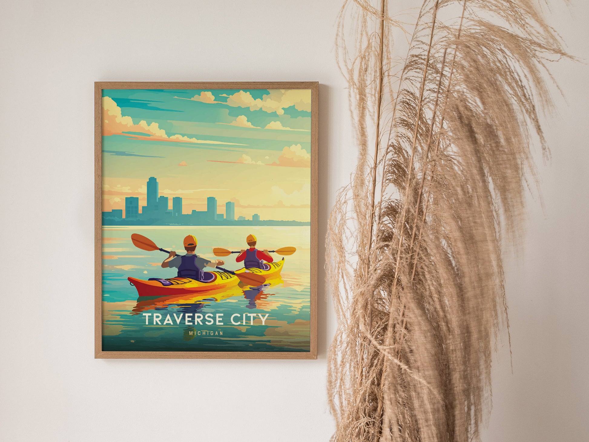 A framed poster of two kayakers on a colorful waterfront, labeled Traverse City, Michigan, hangs on a wall beside tall, dried grasses.