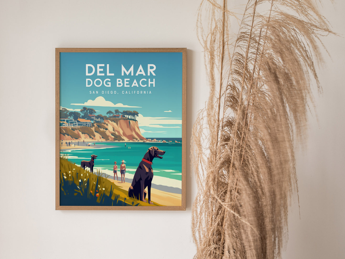 Framed poster of Del Mar Dog Beach showing dogs and people on a sandy beach with cliffs and ocean waves. Tall dried grasses are placed beside the poster.
