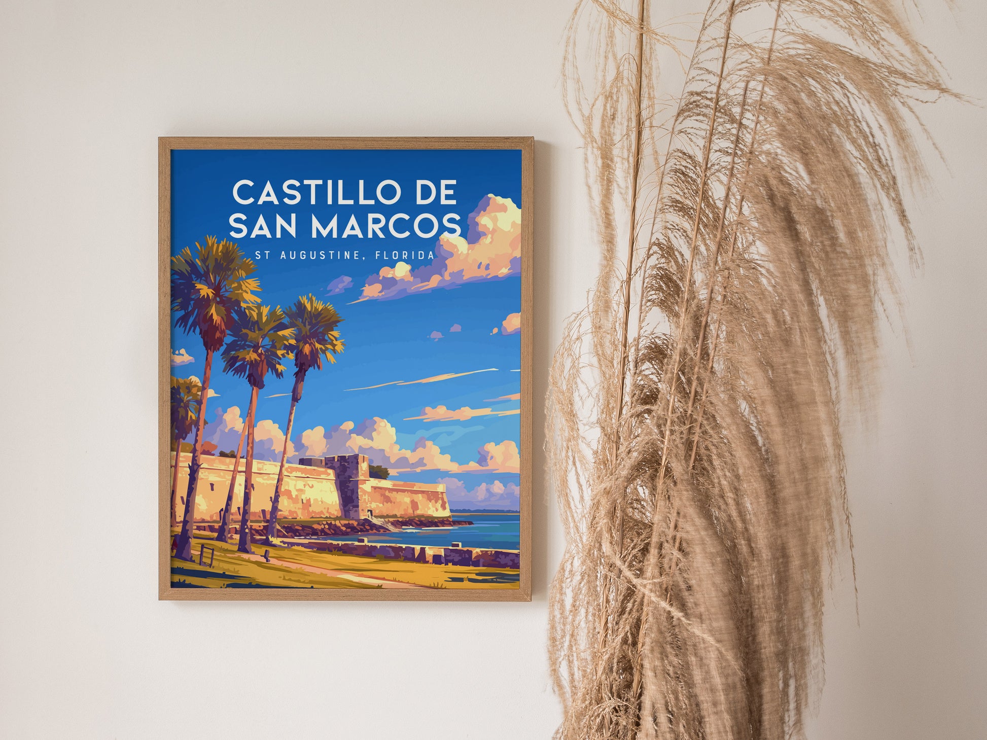 Framed poster of Castillo de San Marcos in St. Augustine, Florida, featuring palm trees and vibrant blue skies. Its displayed on a wall next to decorative dried grasses.