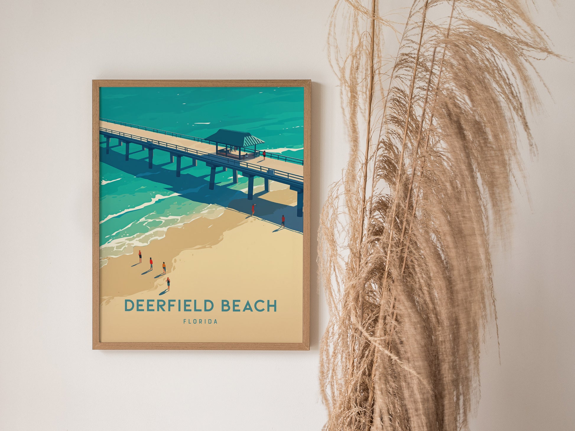 A framed poster of Deerfield Beach, Florida, featuring a pier over turquoise waters and sandy shores, is displayed on a wall next to tall, dried grasses.