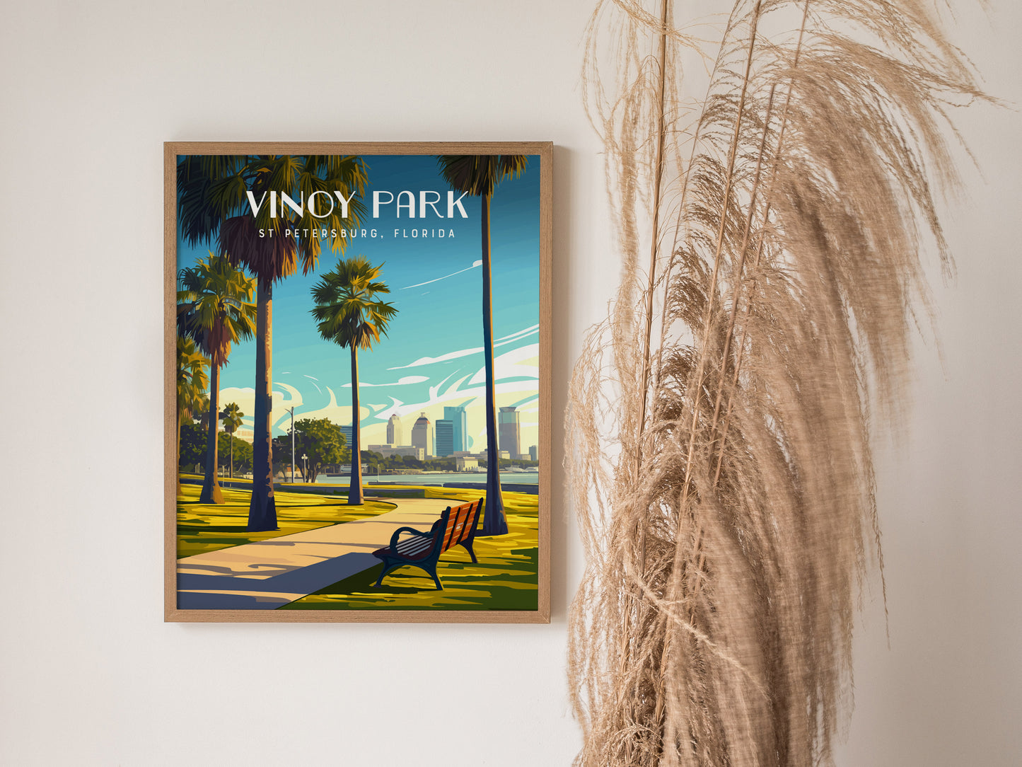 A framed poster of Vinoy Park in St. Petersburg, Florida, featuring palm trees and a waterfront pathway, is displayed on a wall beside dried ornamental grass.