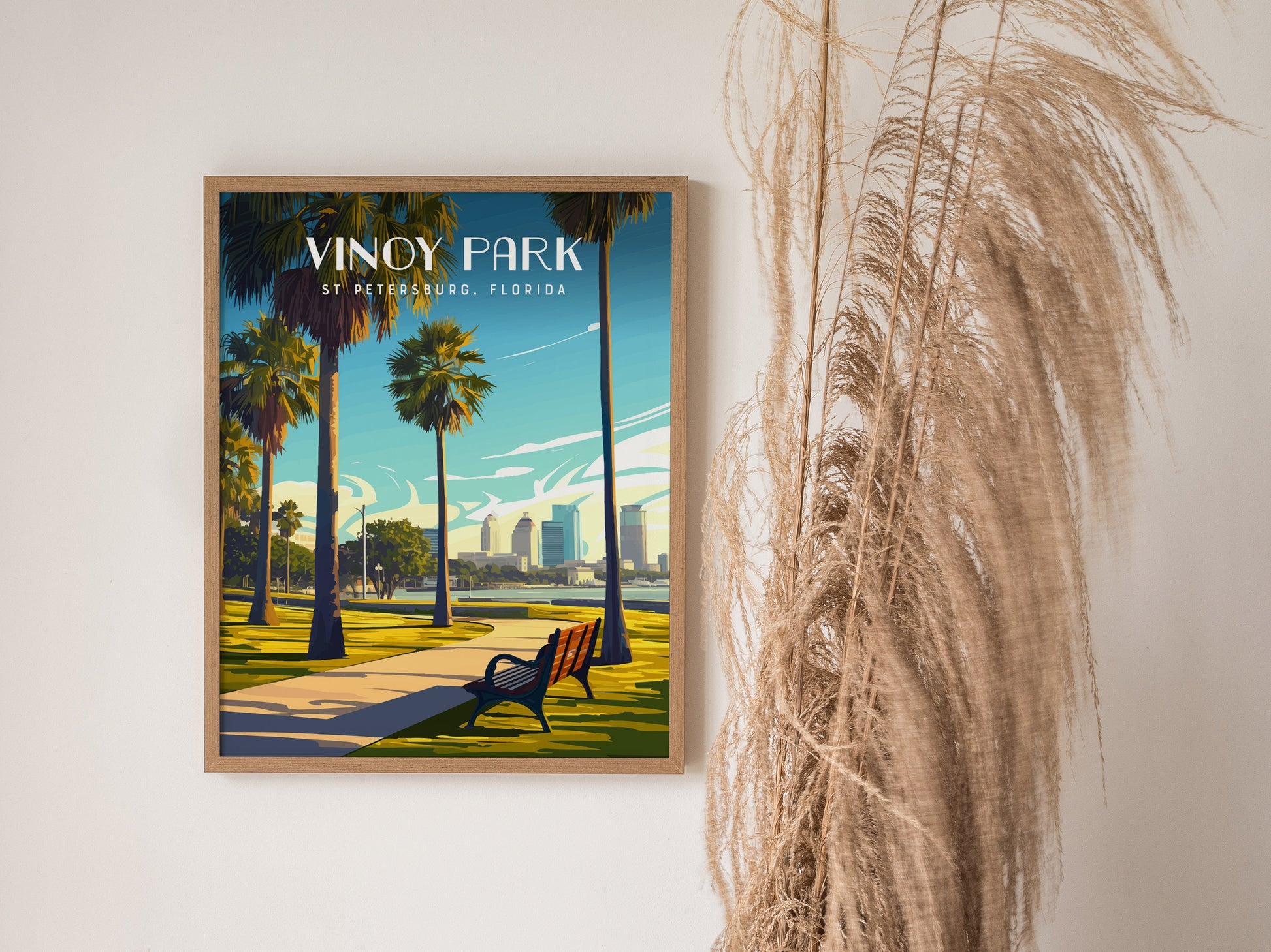 A framed poster of Vinoy Park in St. Petersburg, Florida, featuring palm trees and a waterfront pathway, is displayed on a wall beside dried ornamental grass.