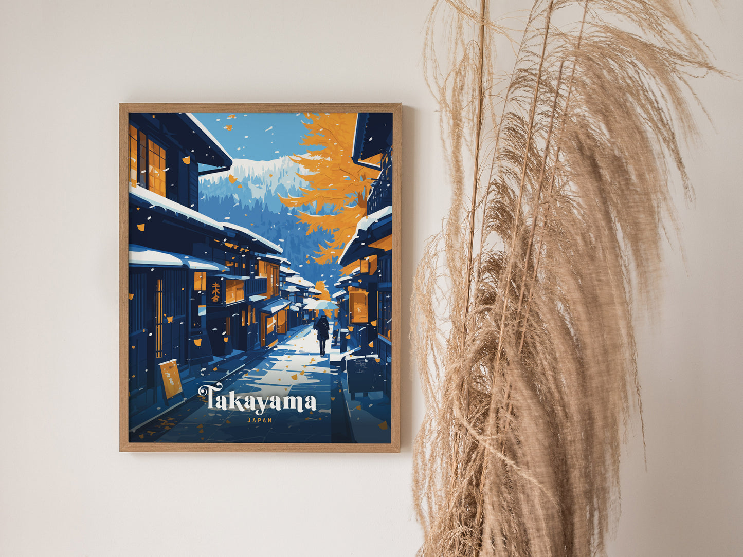 A framed poster of Takayama, Japan, showcasing traditional wooden houses under snow and autumn leaves, with distant mountains. Tall dried grasses are visible beside the artwork.