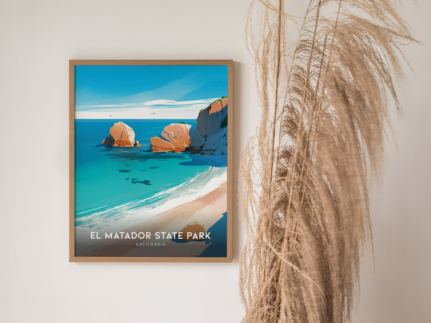 A framed poster of El Matador State Park showcases vibrant blue waters and dramatic cliffs, hung beside tall, wispy pampas grass against a plain wall.