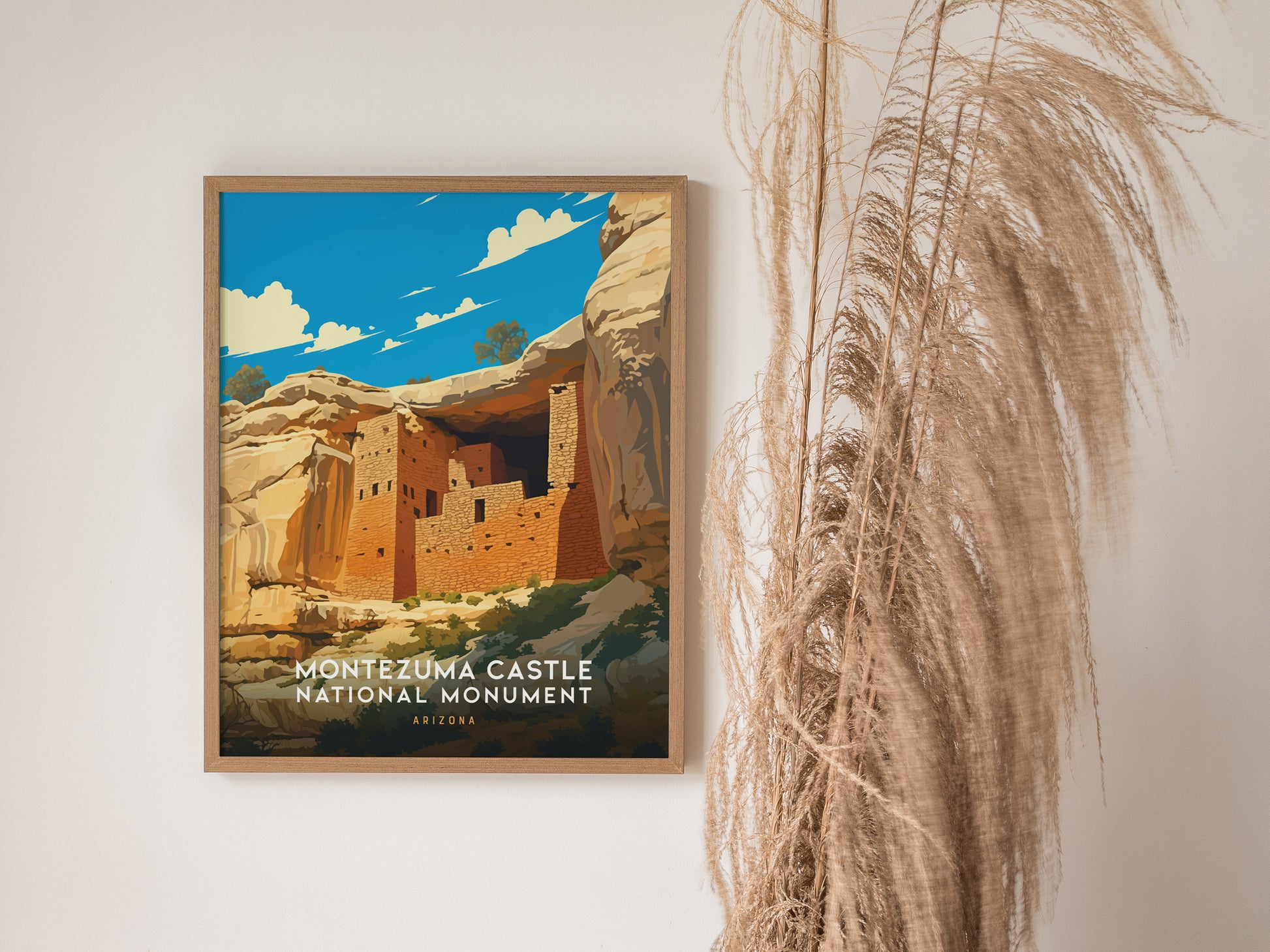 A framed poster of Montezuma Castle National Monument in Arizona hangs on a white wall next to tall, wispy pampas grass. The artwork depicts the cliff dwelling beneath a vibrant.