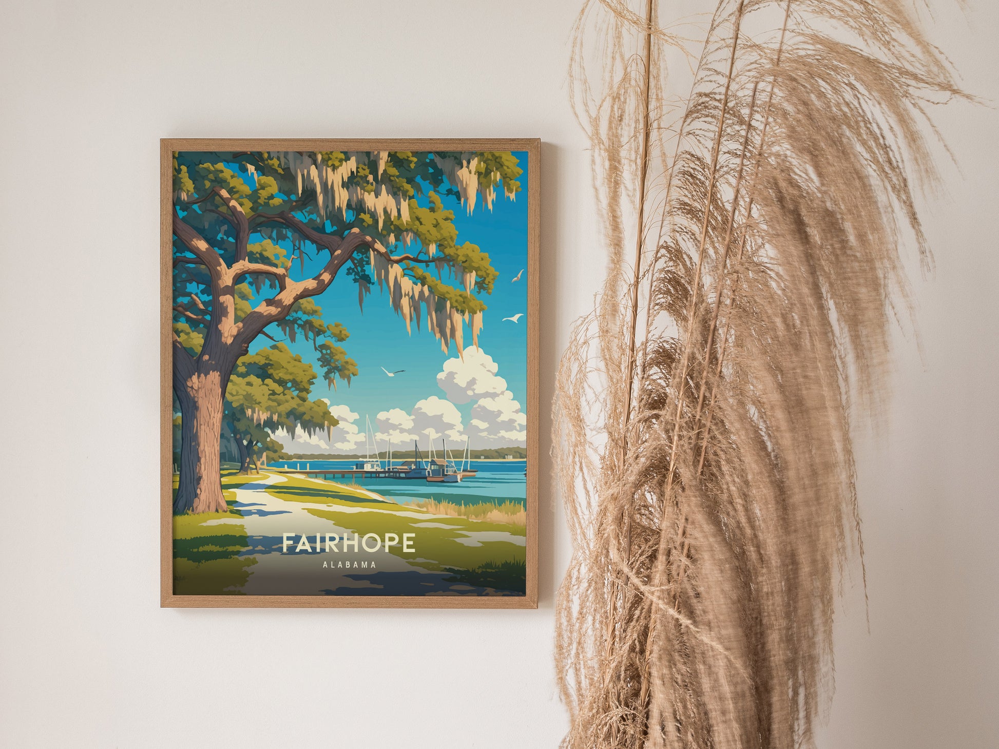 A framed poster of Fairhope, Alabama, featuring a scenic view of a Southern live oak tree and waterfront, hangs on a wall beside decorative dried grasses.