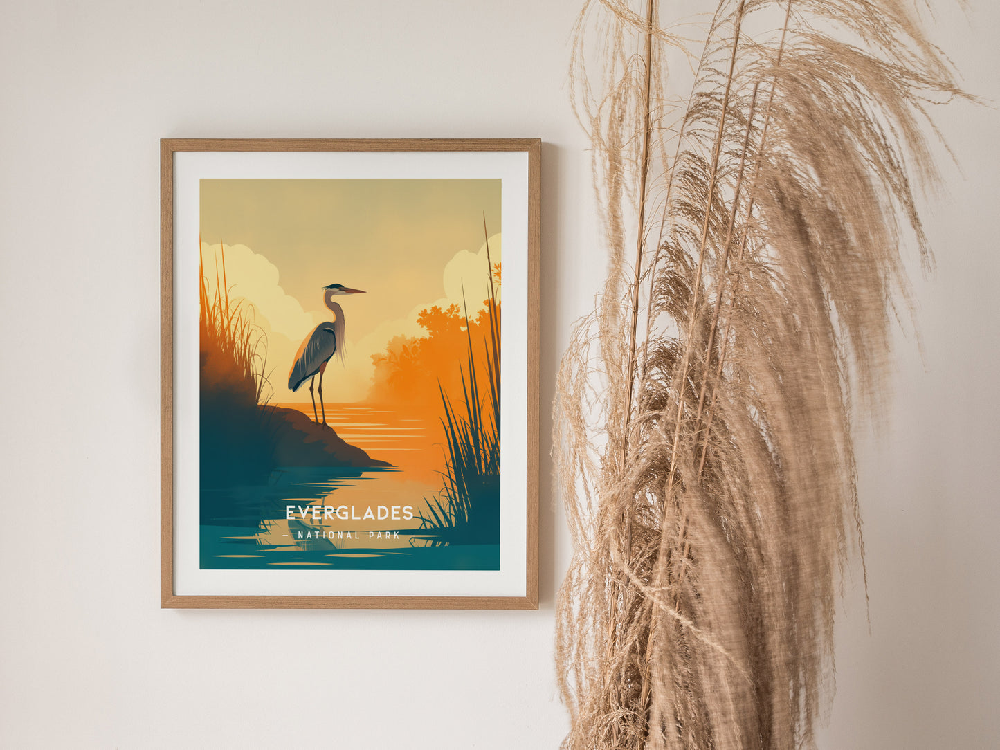 Framed minimalist poster of Everglades National Park featuring a solitary heron by the water at sunset. The artwork is displayed next to tall, dried grasses against a neutral wall.
