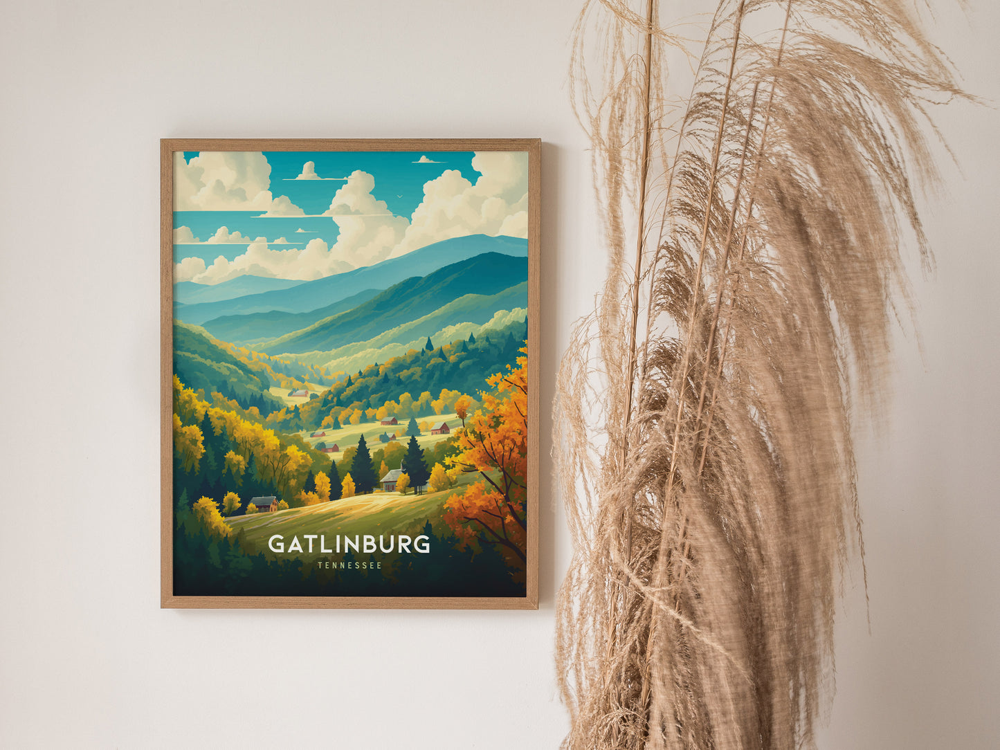A framed Gatlinburg poster depicting lush, colorful landscapes of Tennessees mountains hangs on a wall next to tall, beige pampas grass.