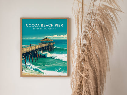 A framed poster of Cocoa Beach Pier hangs on a white wall, next to tall dried grass, showcasing vibrant ocean waves and a thatched-roof pavilion.