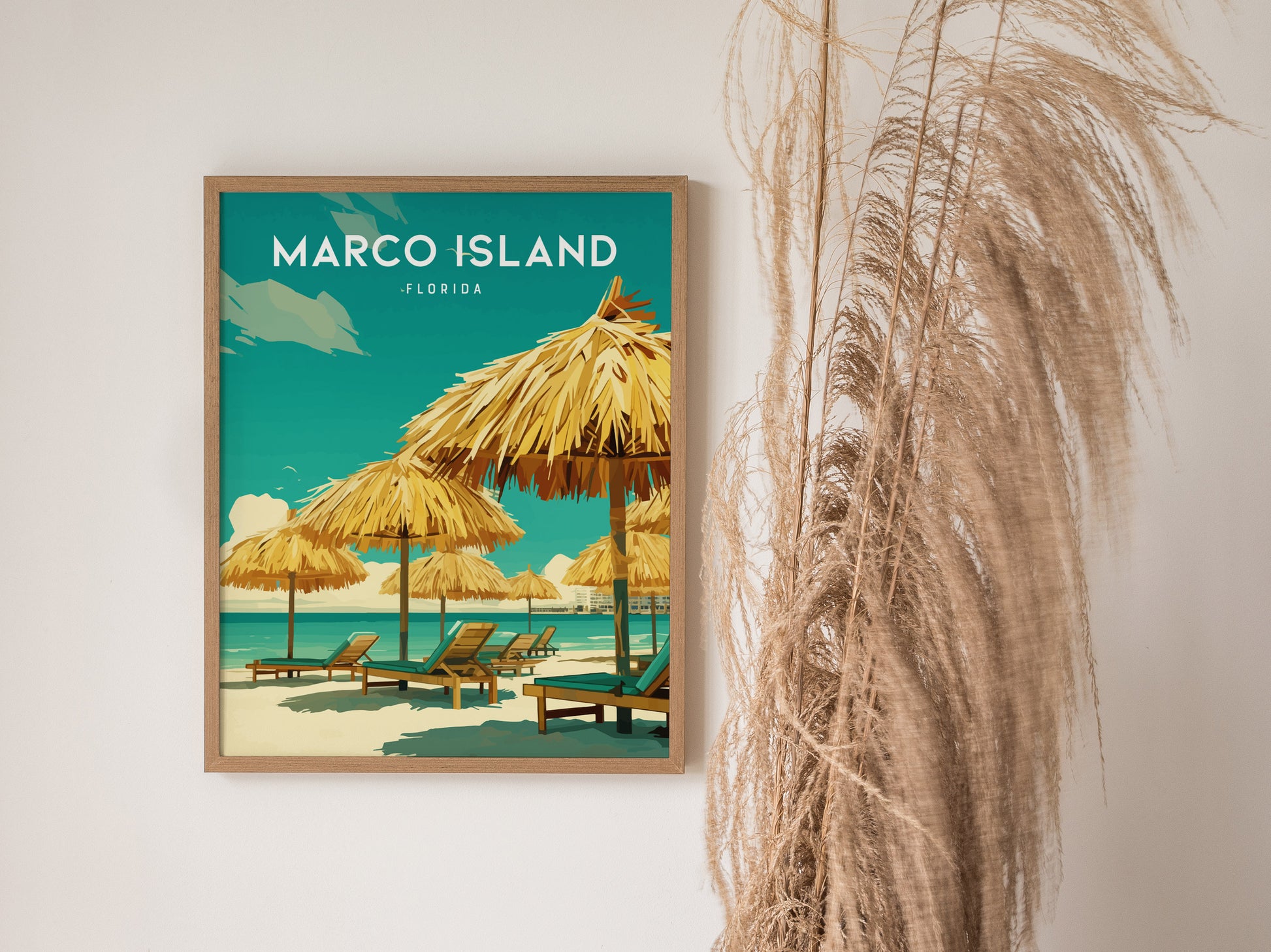 A framed Marco Island, Florida poster features beach chairs and palm thatched umbrellas under a bright sky. Tall, dried grasses are placed beside the poster in a minimalist setting.