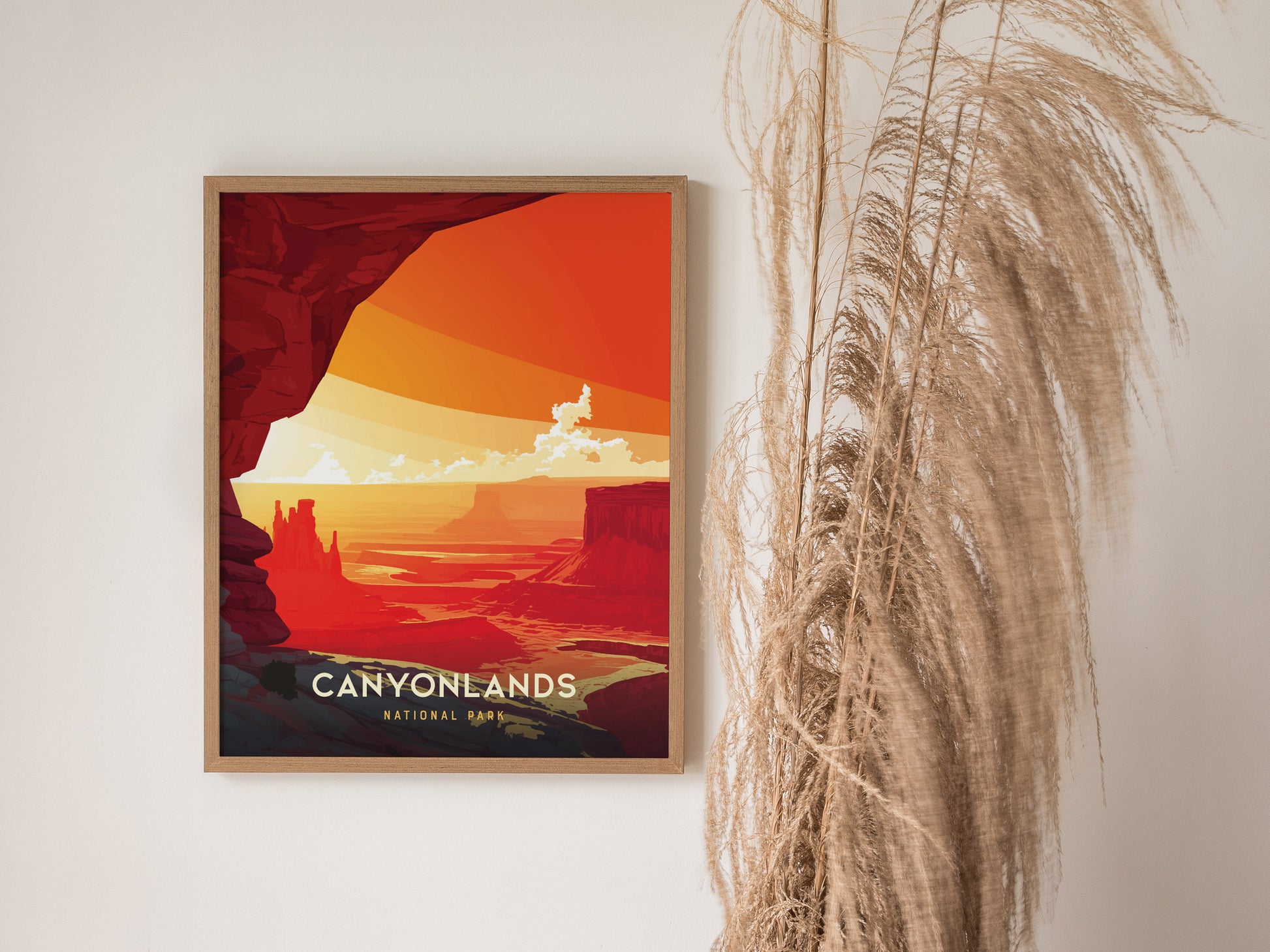 A framed poster of Canyonlands National Park features vibrant orange and red desert canyons, with wispy clouds, set against a white wall and decorative dried grasses.