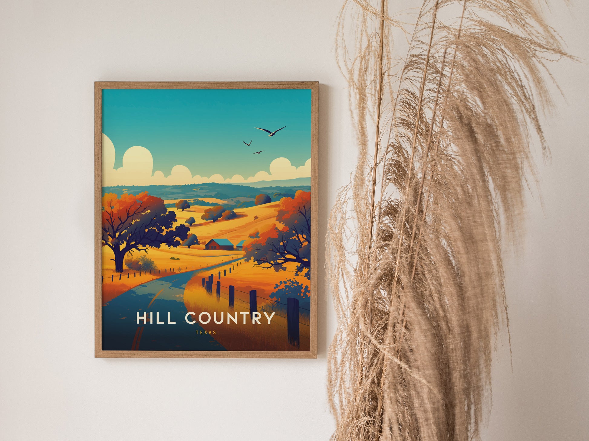 A framed poster depicts a vibrant landscape with rolling hills, colorful trees, and birds flying in the sky, titled Hill Country Texas. Tall dried grasses are placed beside it.