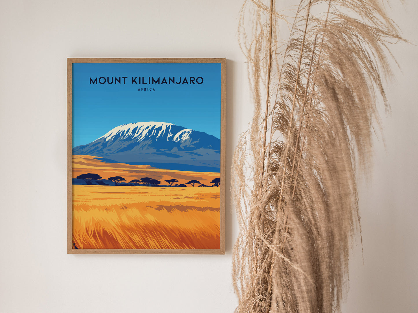 A framed Mount Kilimanjaro poster with a vibrant landscape featuring a snow-capped peak and golden savanna. Dried pampas grass is placed beside the artwork for a decorative touch.