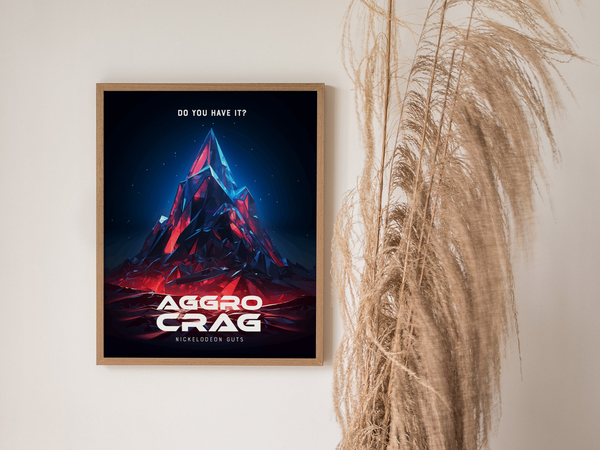 Framed poster featuring a stylized, colorful mountain peak labeled Aggro Crag with Do you have it? text above, next to tall dried grasses against a light wall.