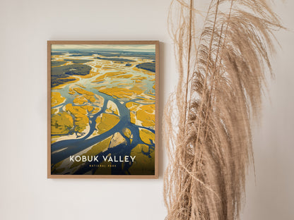 A framed poster of Kobuk Valley National Park features abstract, colorful landscapes with winding rivers and sand dunes. It hangs on a wall beside decorative pampas grass.