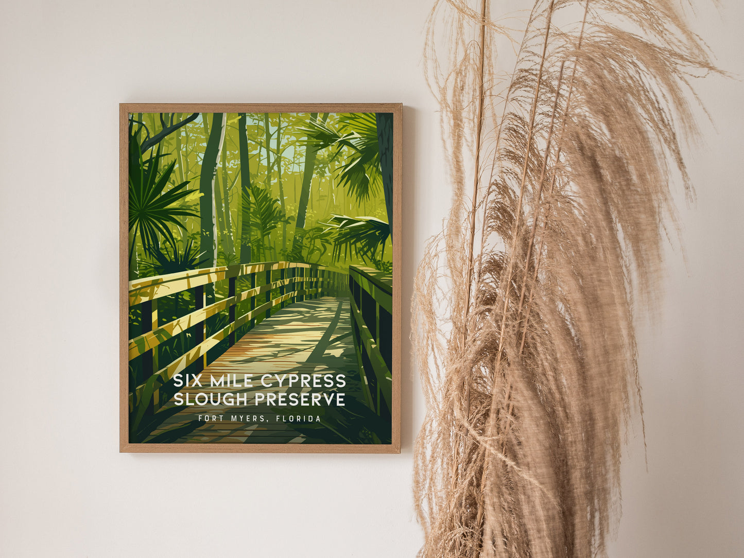 A framed poster of Six Mile Cypress Slough Preserve features a wooden boardwalk surrounded by lush green foliage, displayed against a white wall with dry ornamental grass beside it.