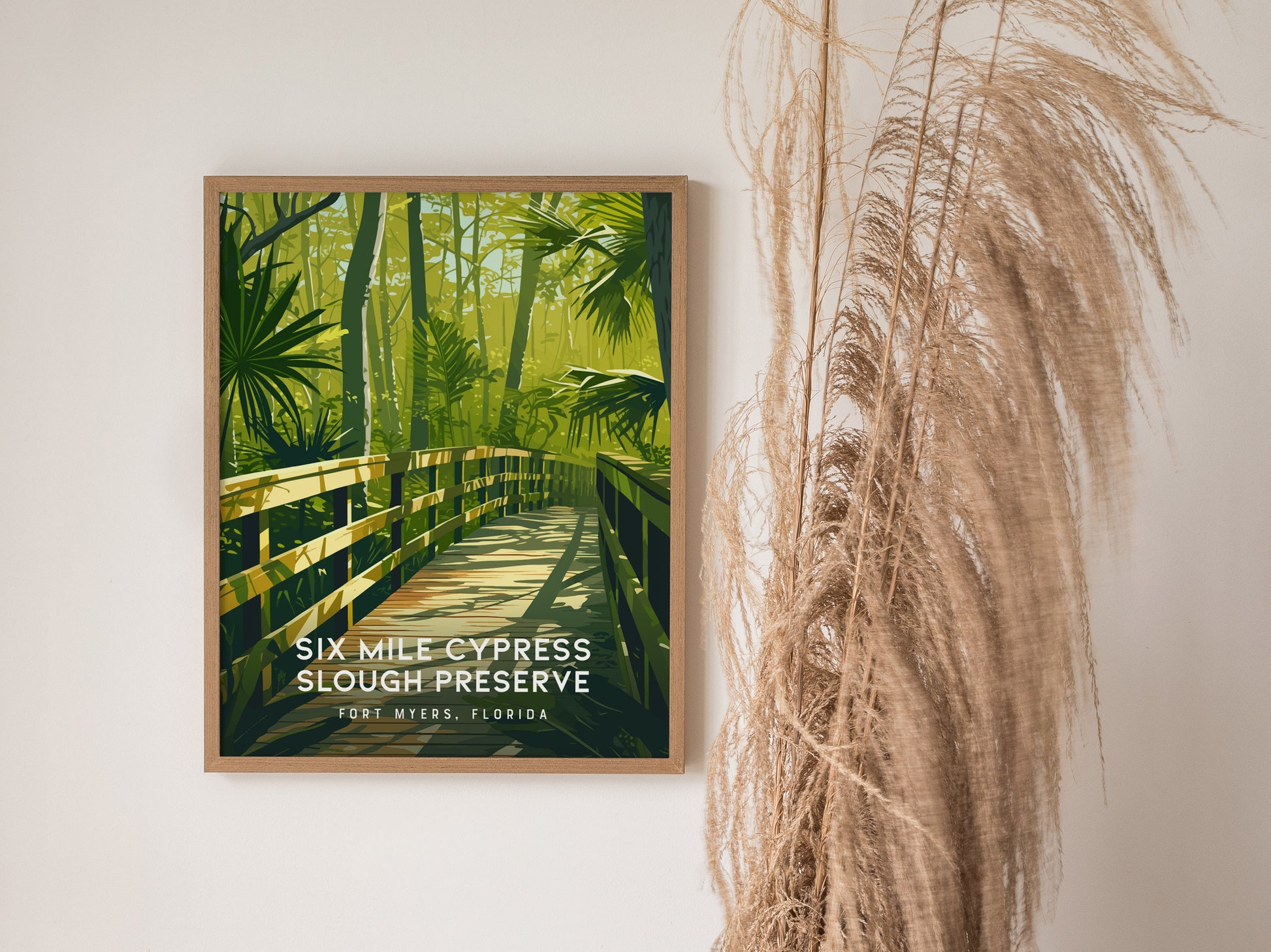 A framed poster of Six Mile Cypress Slough Preserve features a wooden boardwalk surrounded by lush green foliage, displayed against a white wall with dry ornamental grass beside it.