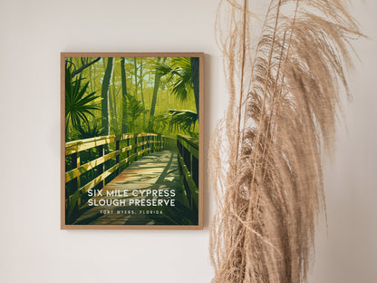 A framed poster of Six Mile Cypress Slough Preserve features a wooden boardwalk surrounded by lush green foliage, displayed against a white wall with dry ornamental grass beside it.