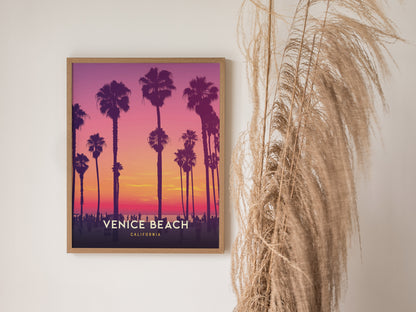 A framed poster of Venice Beach, California, features silhouetted palm trees against a vibrant purple and orange sunset. The poster is displayed on a wall next to dried pampas grass.