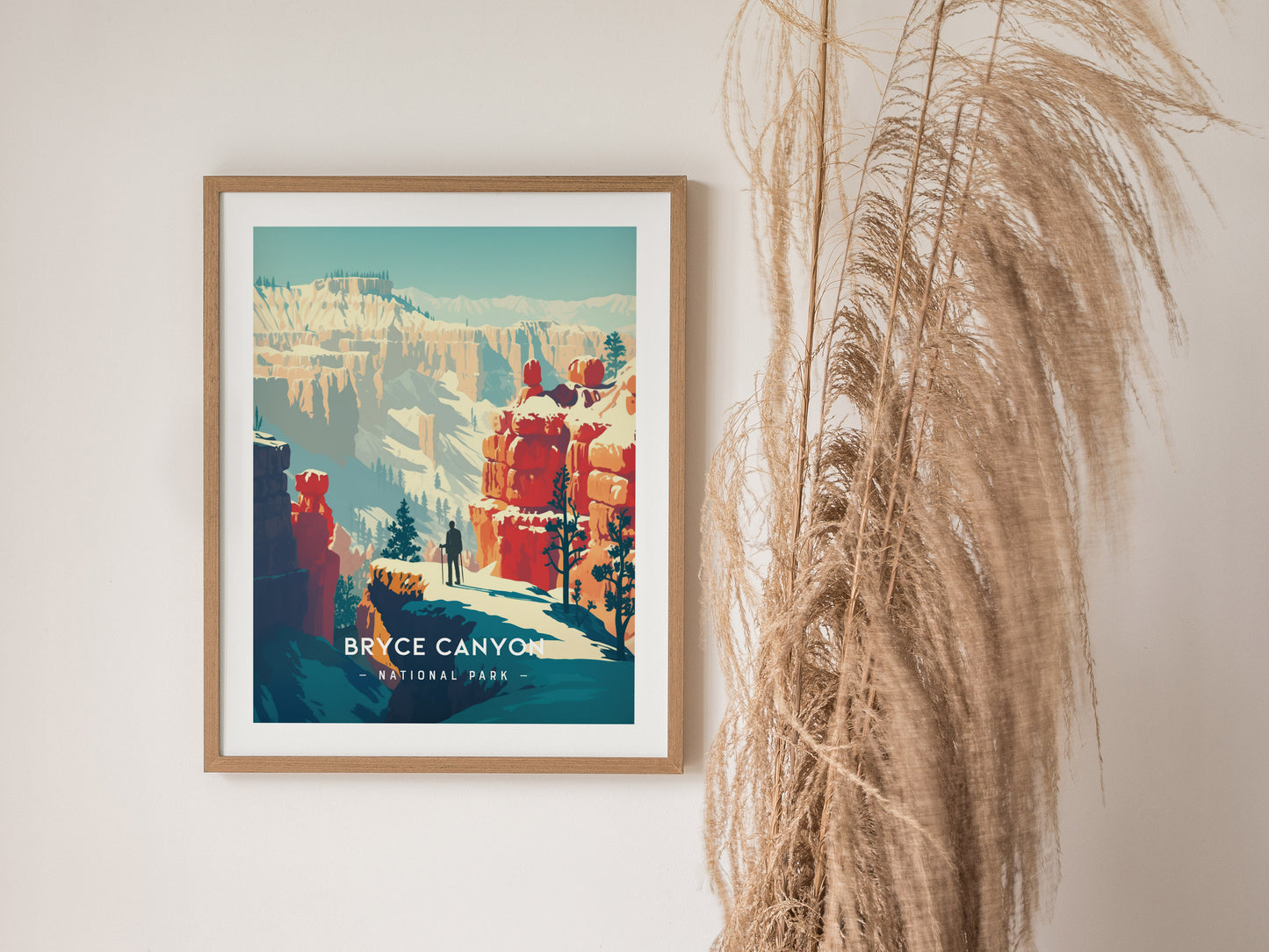 A framed Bryce Canyon National Park print hangs on a white wall, featuring vibrant red rock formations and a figure standing on a cliff. Pampas grass decoratively leans beside it.